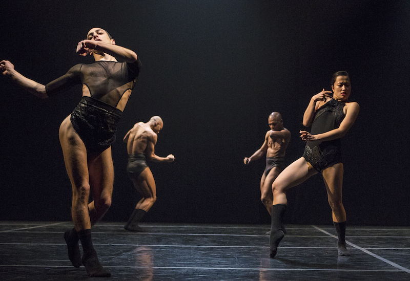 Dancers in black briefs and leotards sway the hips and gesticulate. Two men in the background stand with legs together.